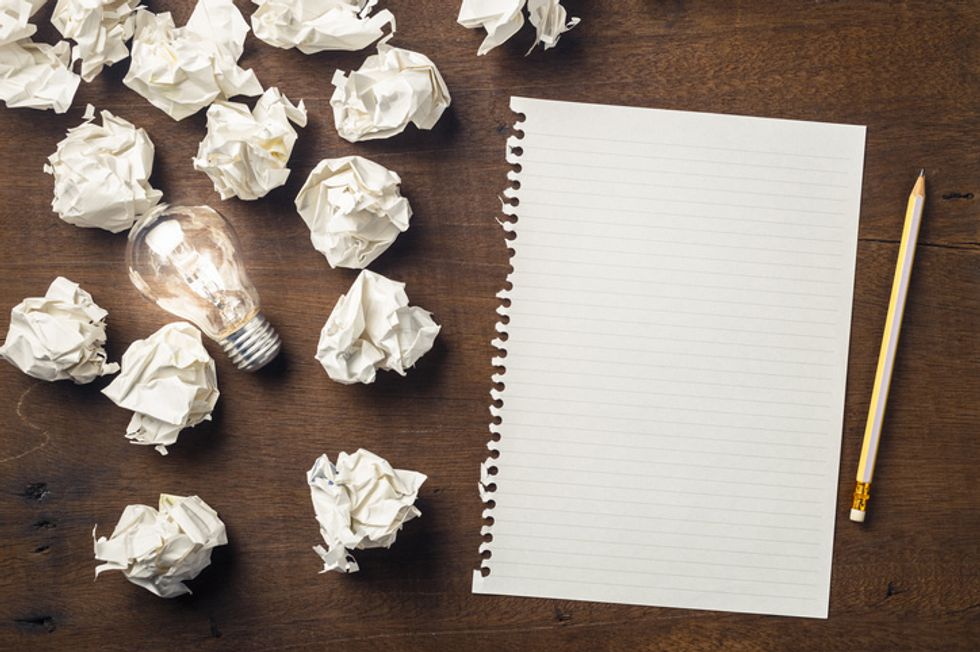 a wood table with crumpled up paper balls next to a blank sheet of paper and a pencil. There is a lightbulb amongst the paper.