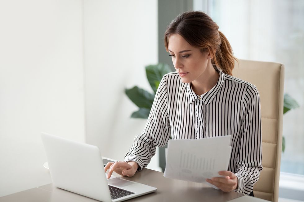 Serious focused businesswoman typing on laptop holding papers in a remote work setting