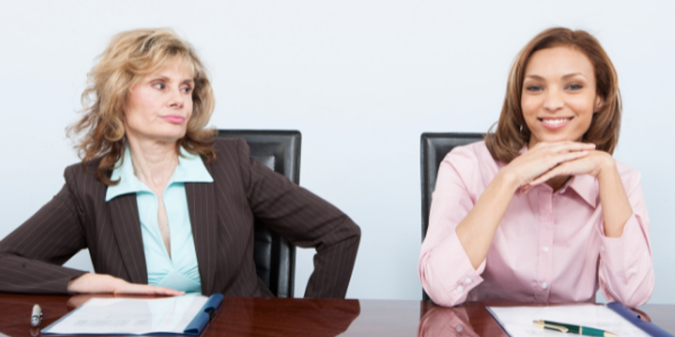 Two female coworkers; woman on left looking awkwardly at woman on the right, who is smiling